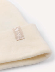A close-up of the Beanie – Merino Baby AirWool, a cream-colored knit baby beanie crafted from merino wool. It features a small beige tag with white text and logo, has a folded edge, and is displayed on a white background to showcase its breathable insulation.