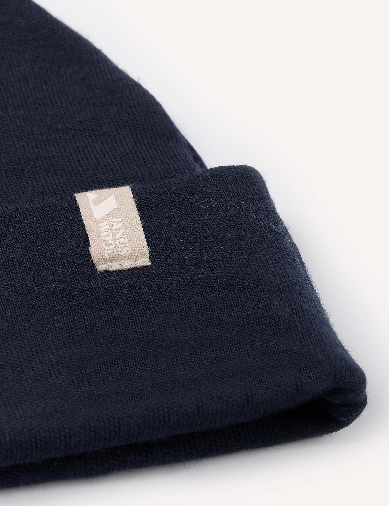 Close-up of the Beanie – Merino Baby AirWool in navy blue, crafted from merino wool, featuring a beige rectangular tag with a simple logo and text sewn onto the cuff, set against a plain white background.