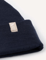 Close-up of the Beanie – Merino Baby AirWool in navy blue, crafted from merino wool, featuring a beige rectangular tag with a simple logo and text sewn onto the cuff, set against a plain white background.