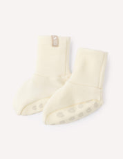 A pair of Booties – Merino Baby AirWool in a cream color, featuring non-slip paw print patterns on the soles, displayed on a white background.