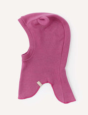 The Balaclava – Merino Baby AirWool is a pink, hooded balaclava crafted from soft merino wool, shown laid flat on a white background. It features a curved face opening and an extended neck section, with a small beige tag sewn onto the side.