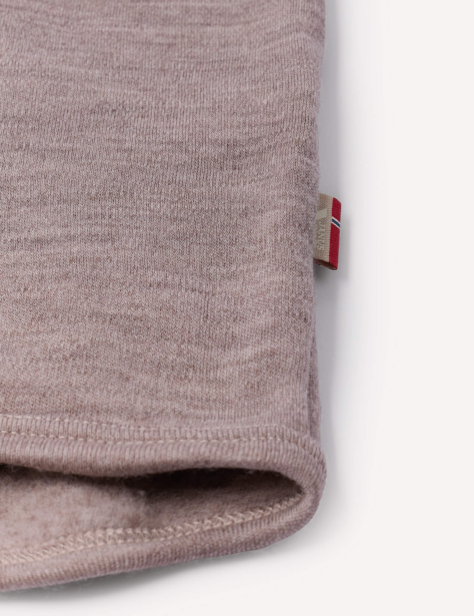 Close-up of a folded edge of a soft, light brown fabric from the Jacket – Merino Baby AirWool, with a small tag featuring a red and white design sewn into the seam. The background is plain and light-colored.