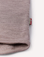 Close-up of a folded edge of a soft, light brown fabric from the Jacket – Merino Baby AirWool, with a small tag featuring a red and white design sewn into the seam. The background is plain and light-colored.