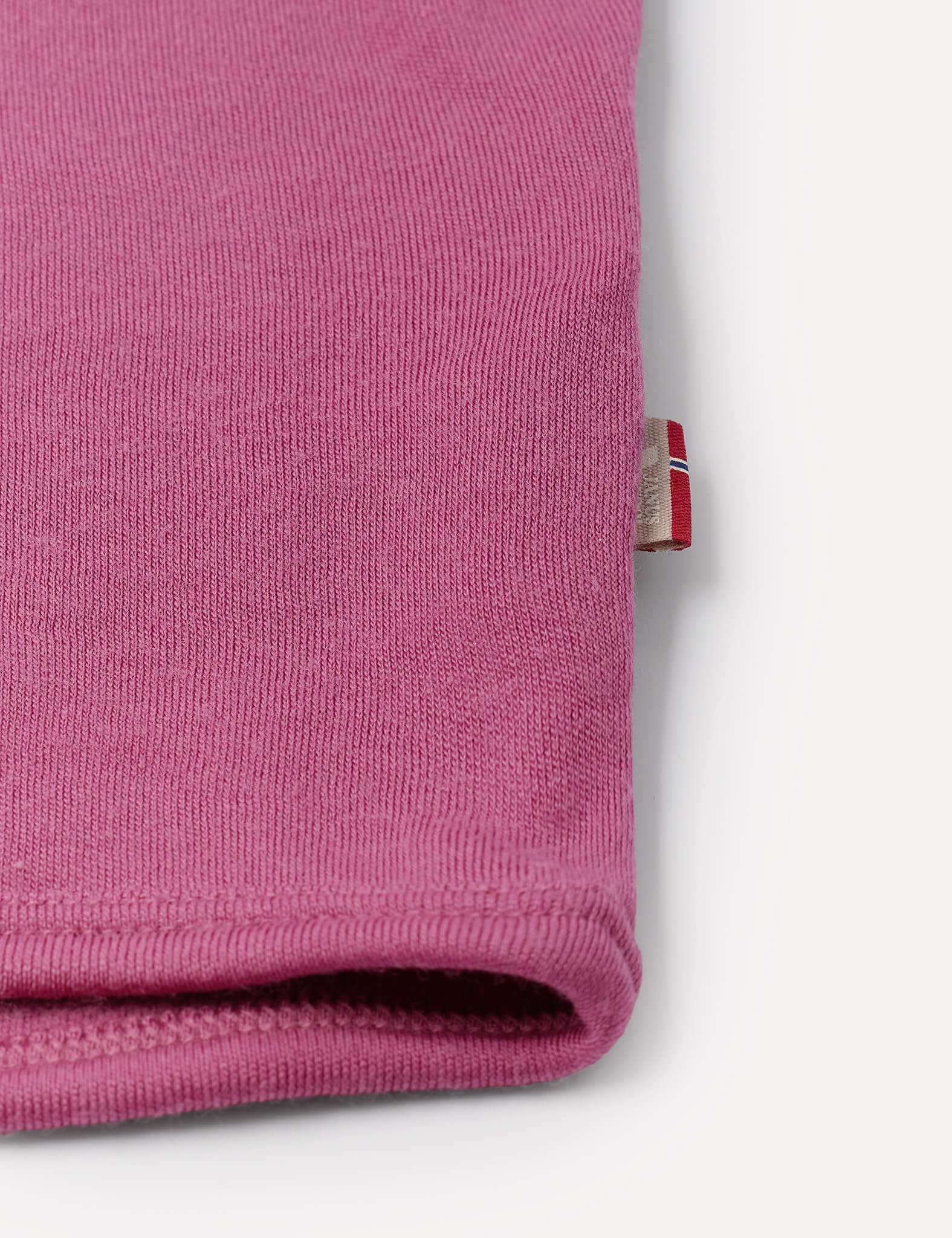 Close-up of the hem of the pink ribbed fabric Jacket – Merino Baby AirWool, possibly a sleeve or pant leg, with a small beige tag featuring red and white stripes visible at the edge. The soft material suggests features like breathable insulation. The background is white.
