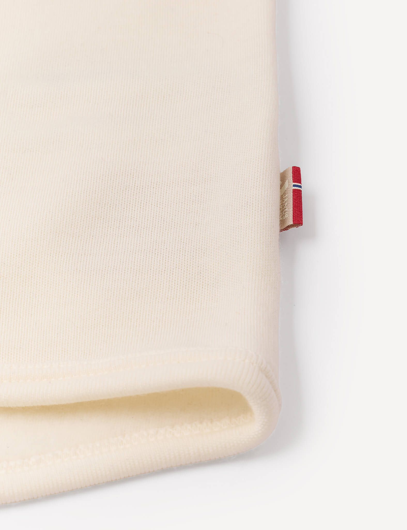 A close-up of a cream-colored folded fabric from the Jacket – Merino Baby AirWool, featuring a small red, white, and blue tag sewn into the seam on a white background, showcasing the breathable insulation typical of this high-quality merino wool jacket.