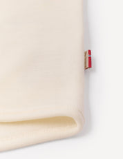 A close-up of a cream-colored folded fabric from the Jacket – Merino Baby AirWool, featuring a small red, white, and blue tag sewn into the seam on a white background, showcasing the breathable insulation typical of this high-quality merino wool jacket.