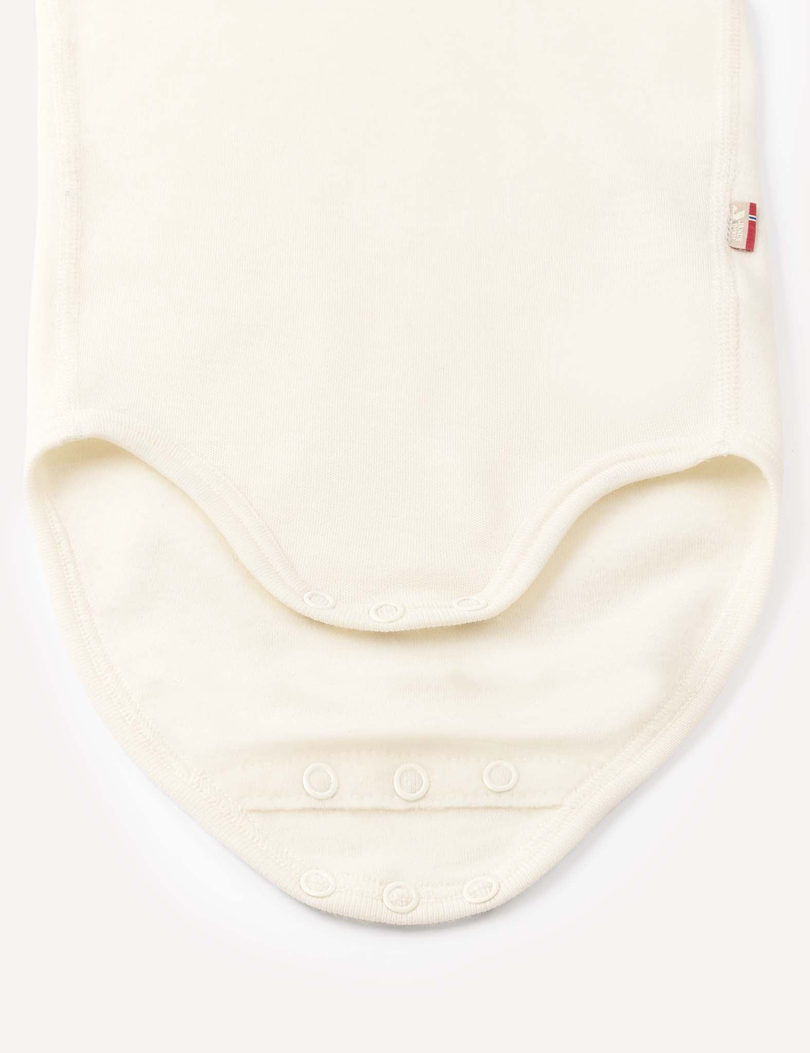 The Body – Merino Baby AirWool is displayed flat in a cream color, highlighting the bottom area with three metal snap buttons along the curved edge for easy fastening. Crafted from soft merino wool, this bodysuit ensures breathable comfort, and a small red fabric tag appears on the right side.