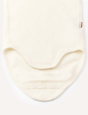 The Body – Merino Baby AirWool is displayed flat in a cream color, highlighting the bottom area with three metal snap buttons along the curved edge for easy fastening. Crafted from soft merino wool, this bodysuit ensures breathable comfort, and a small red fabric tag appears on the right side.
