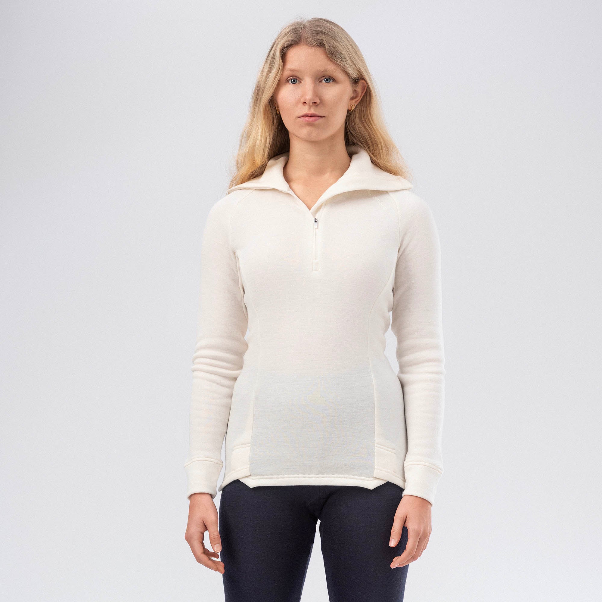 A woman with long blonde hair stands against a light background, wearing the Polo Zip Merino – AirWool 260 Women in Marshmallow and dark pants, facing forward with a neutral expression to highlight the pullover's warmth and breathability.