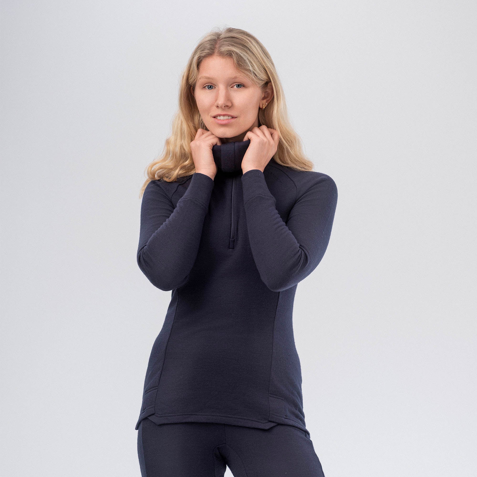 A woman with long blonde hair wears the Polo Zip Merino – AirWool 260 Women in navy, paired with matching pants. She stands against a light background, holding the collar and looking at the camera with a slight smile.