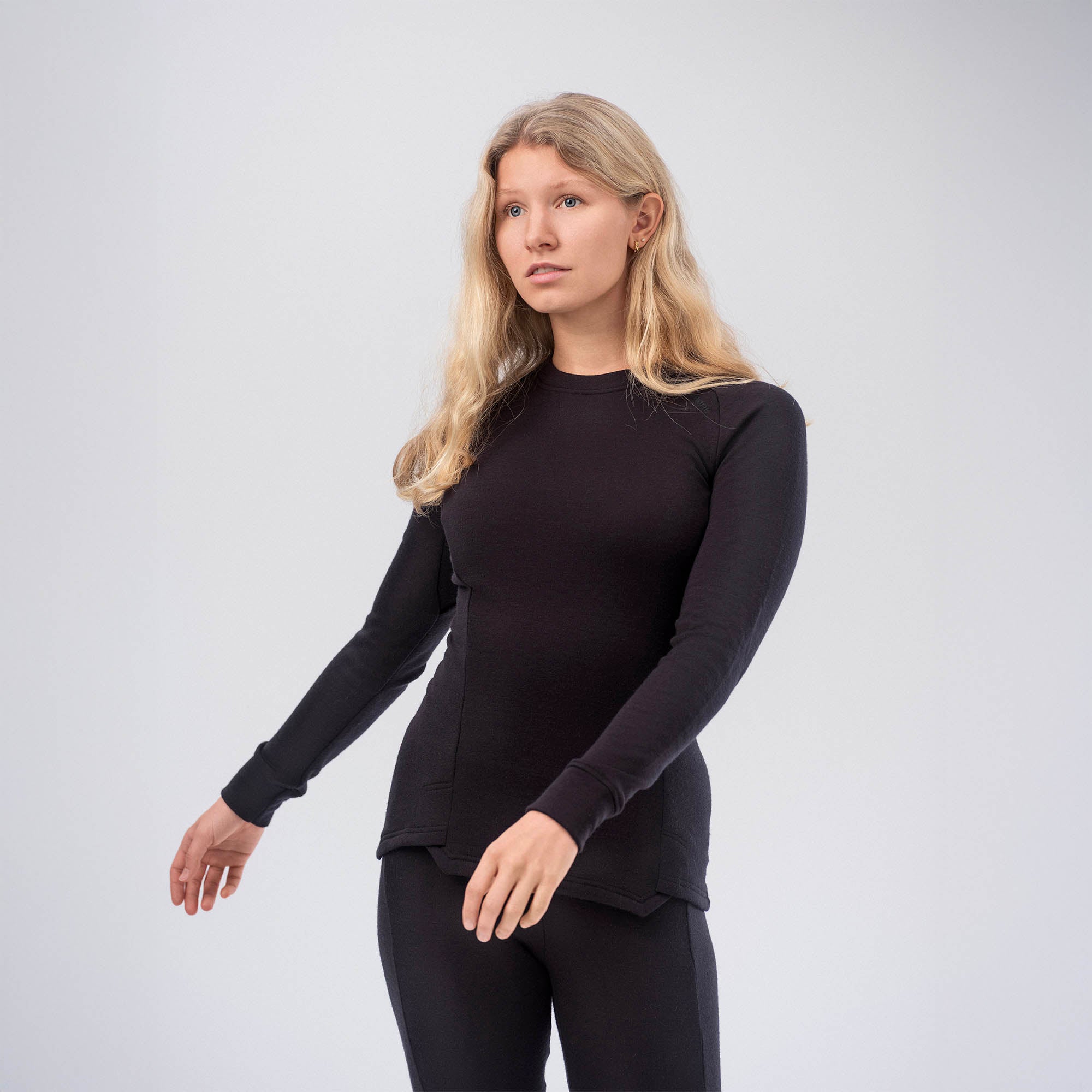 A woman with long blonde hair wears a fitted black Jersey Merino – AirWool 260 Women, standing against a plain light background and looking to the side, showcasing the warmth and breathability of the jersey.
