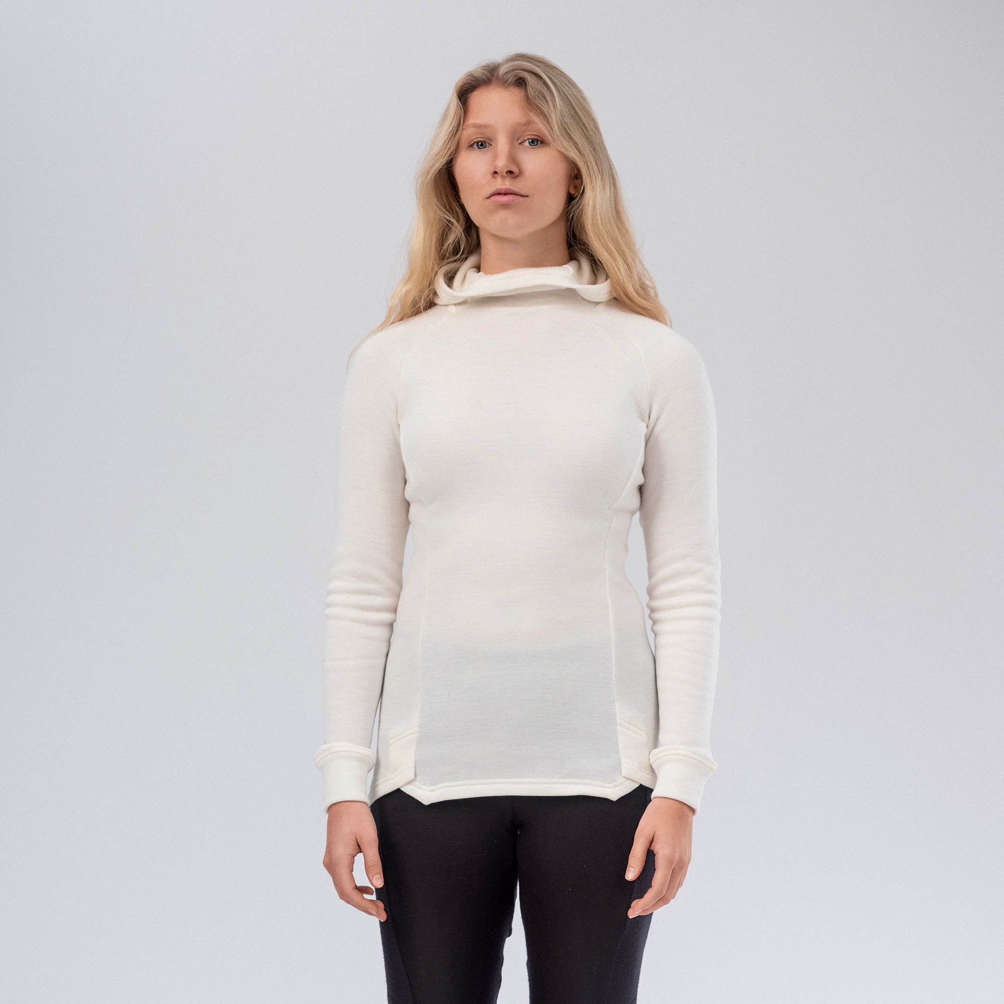 A woman with long blonde hair wears the Marshmallow-colored Hooded Jersey Merino – AirWool 260 Women sweater and black pants, standing against a plain light gray background with a neutral expression.