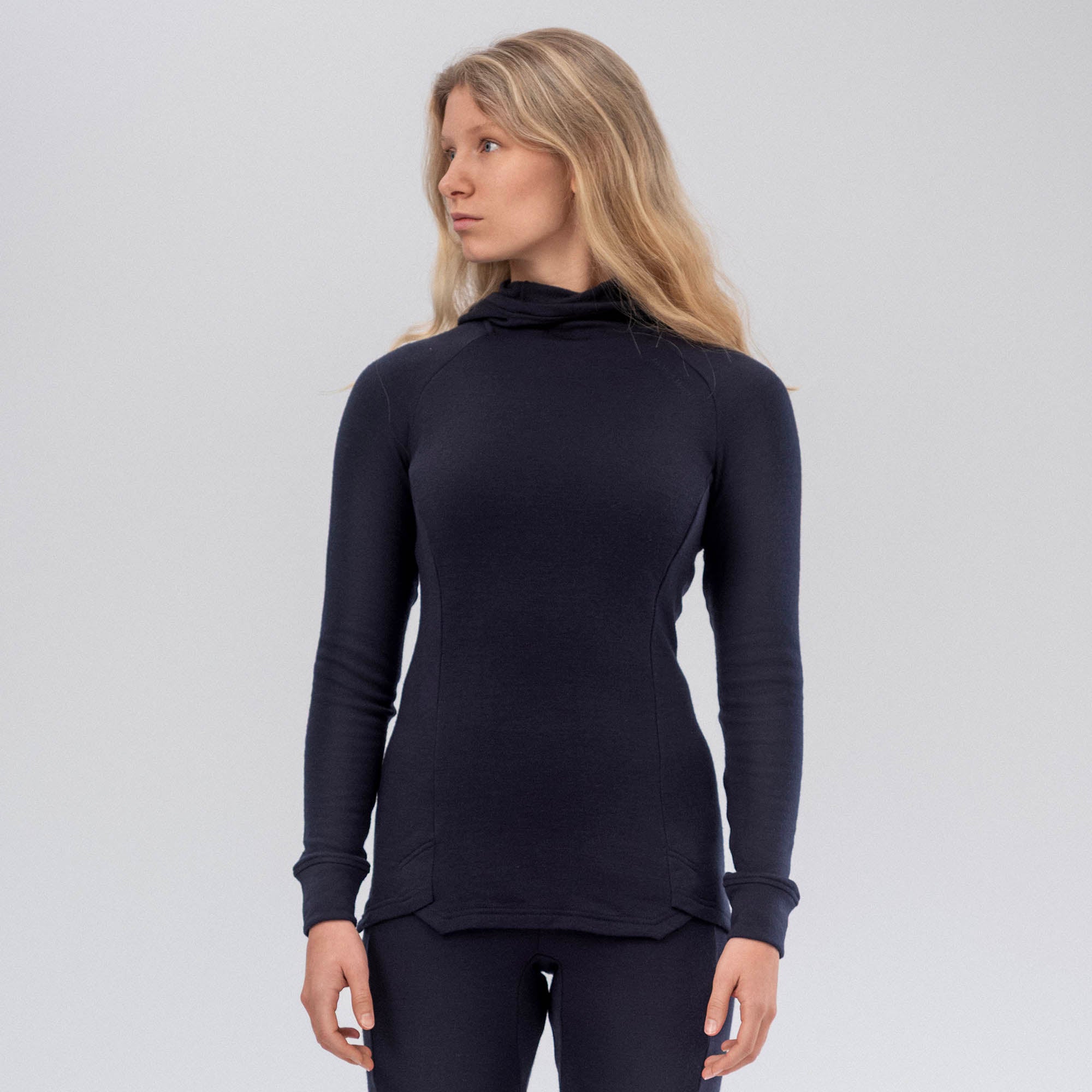A woman with long blonde hair, wearing the AirWool 260 Women Hooded Jersey Merino set in dark navy, stands facing slightly left against a plain light gray background.