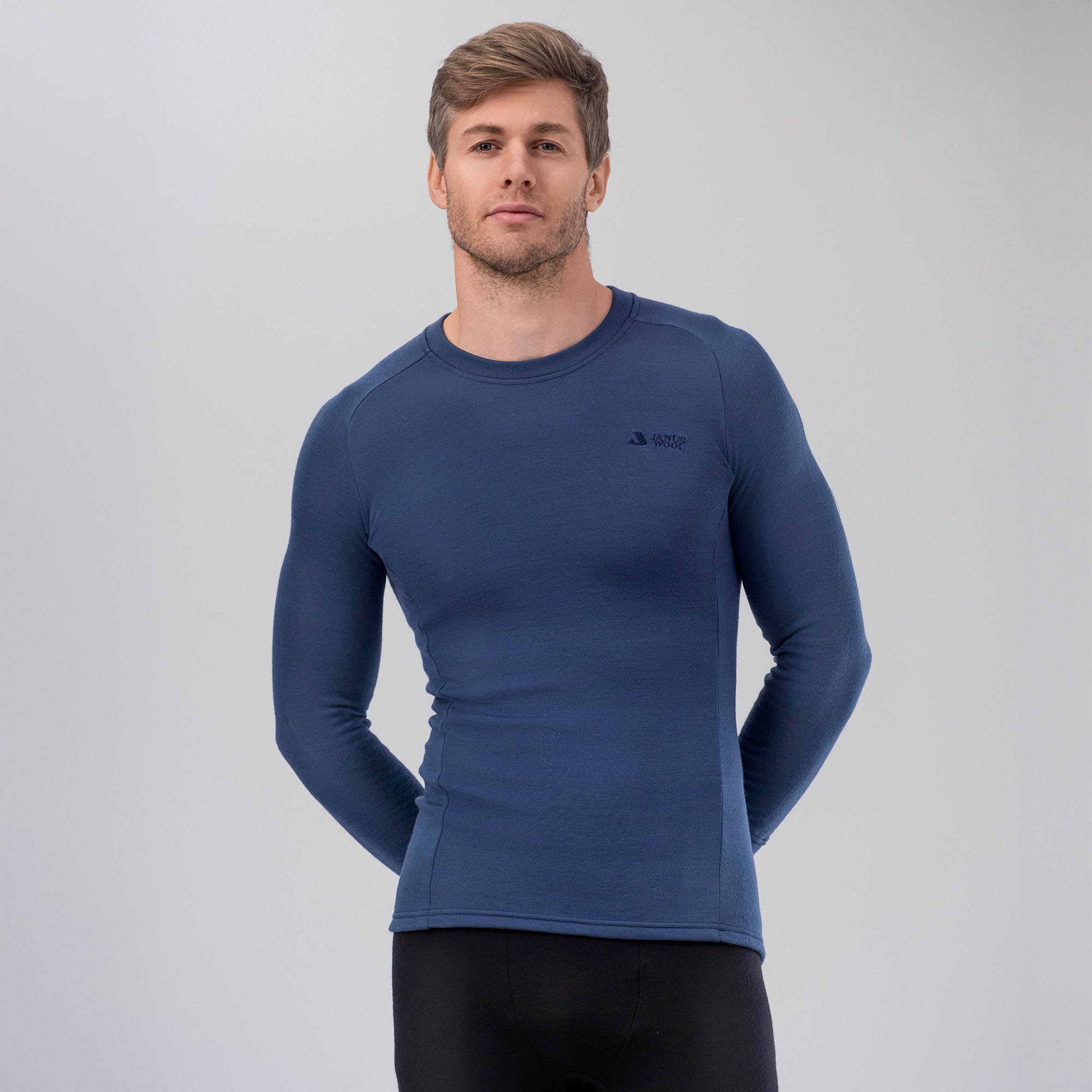 A man stands against a light gray background, wearing the Jersey Merino – AirWool 260 Men in navy blue and black pants. The fitted long-sleeve shirt provides warmth and breathability. His hands are behind his back as he looks ahead with a neutral expression.