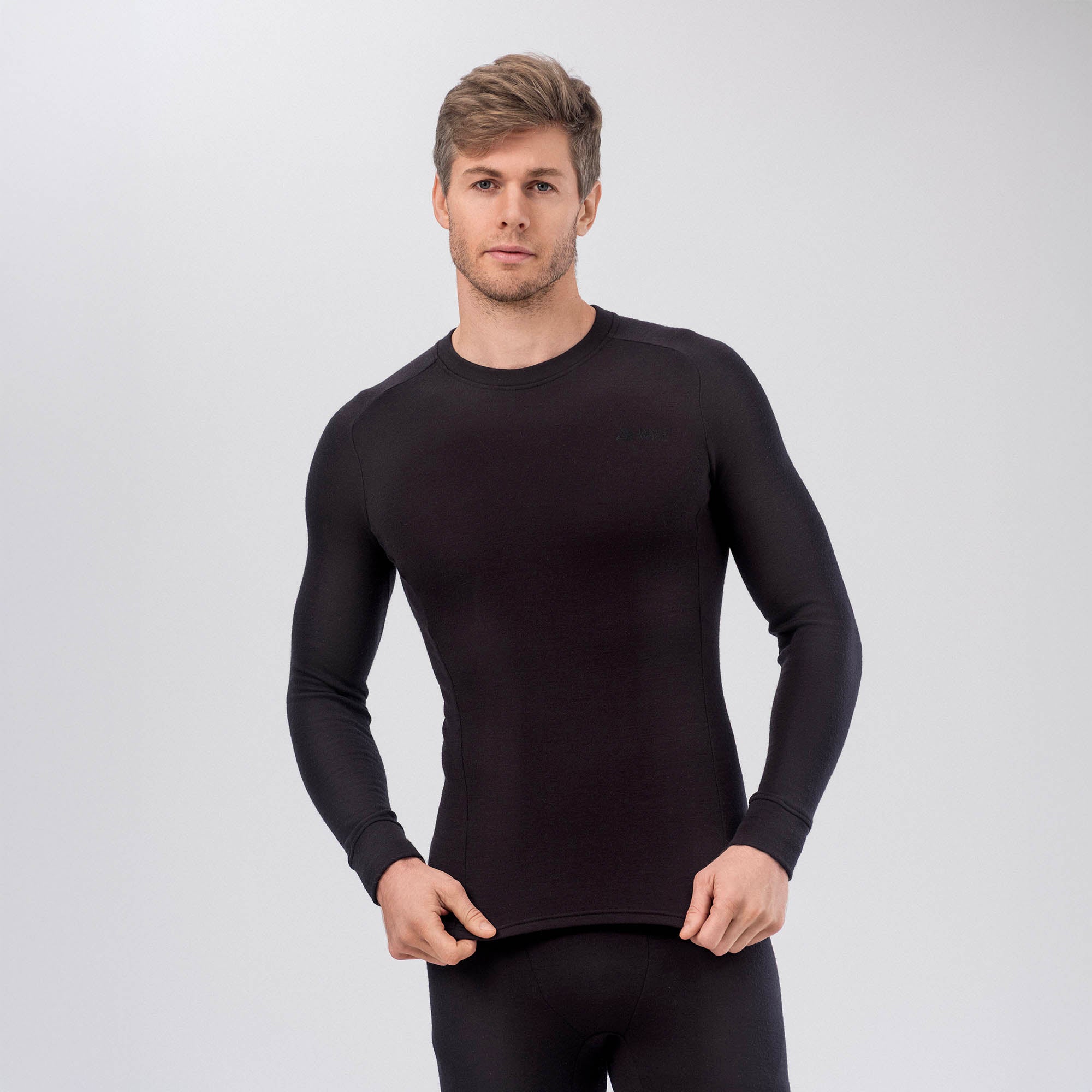 A man with short light brown hair wears the Jersey Merino – AirWool 260 Men long-sleeve shirt and matching pants, standing against a light gray background. Holding the hem, he highlights the product’s warmth and breathability.