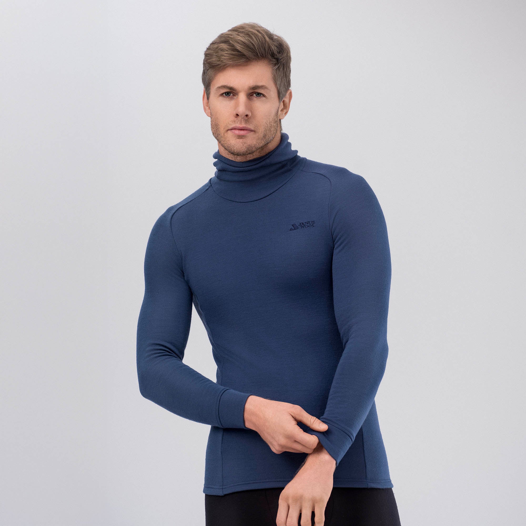 A man with short light brown hair wears the Hooded Jersey Merino – AirWool 260 Men in Ensign Blue and black pants, standing against a plain light grey background and looking straight at the camera.