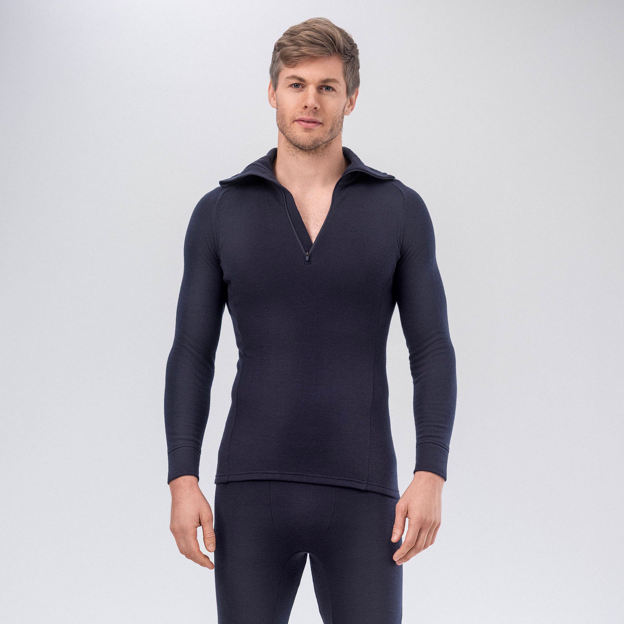 A man stands facing forward, wearing a fitted, long-sleeve, dark-colored merino wool thermal top with a half-zip collar and matching thermal bottoms against a plain light background.