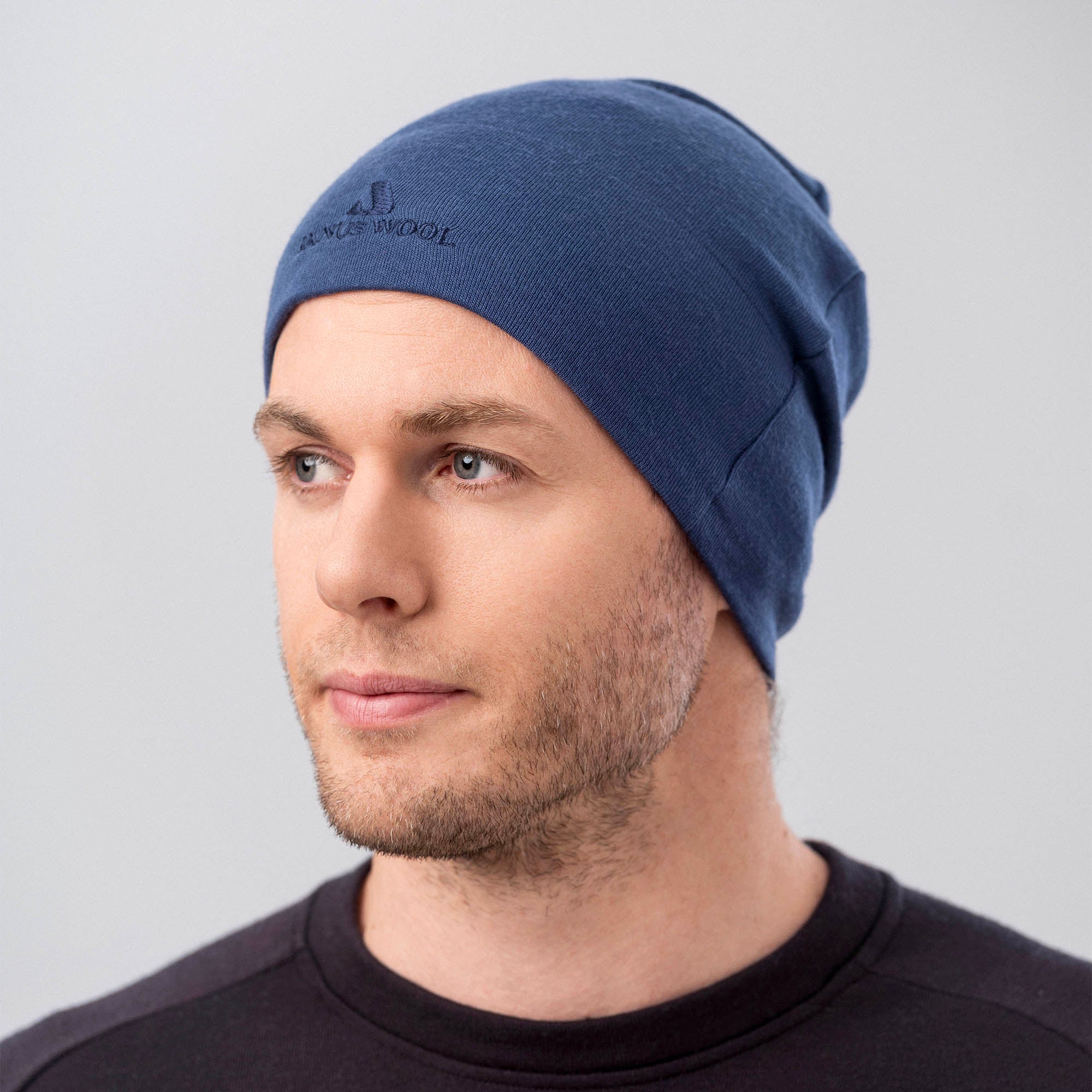 A man wearing the Beanie Merino – Airwool 260, a blue merino wool beanie known for warmth and breathability, and a black shirt looks slightly to the side against a light gray background.