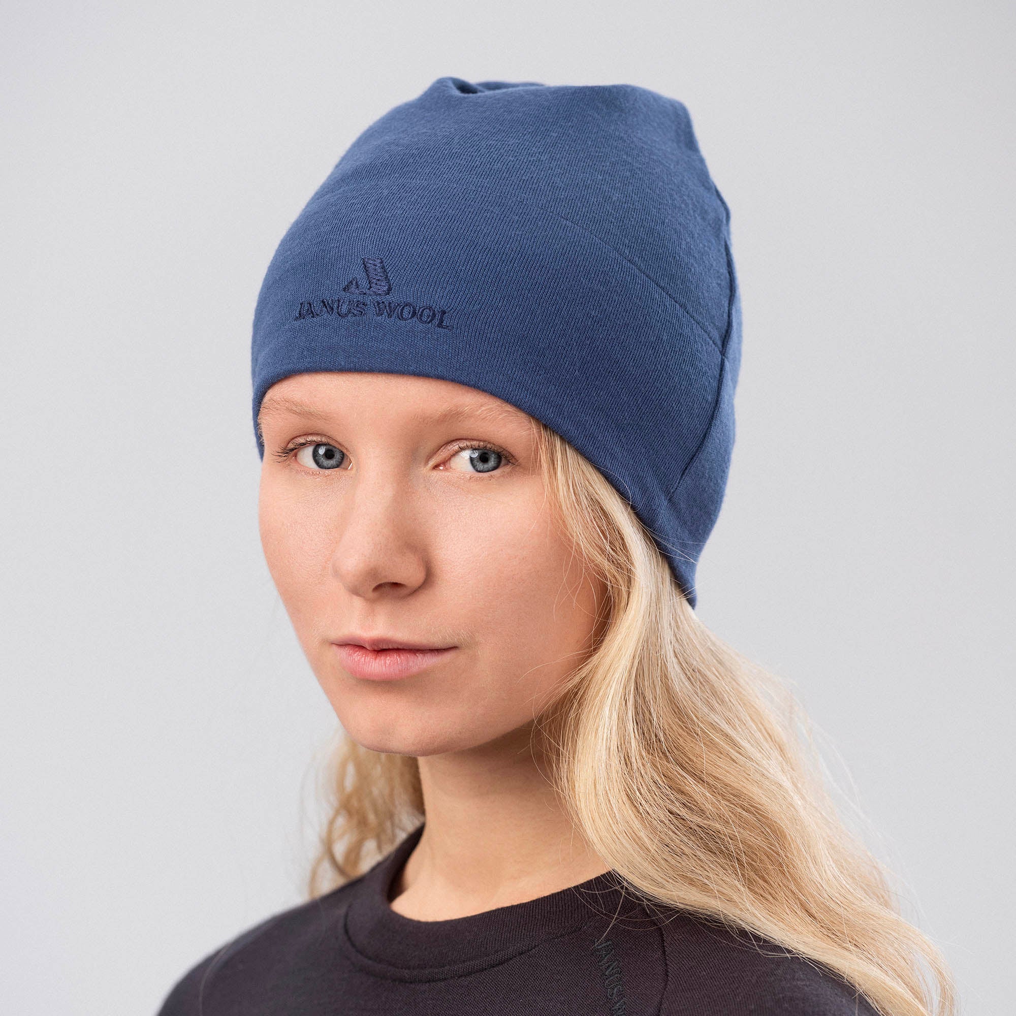 A person with long blonde hair wears the Beanie Merino – Airwool 260 in Ensign Blue and a dark top, facing the camera with a neutral expression. The beanie provides warmth against a plain light background.