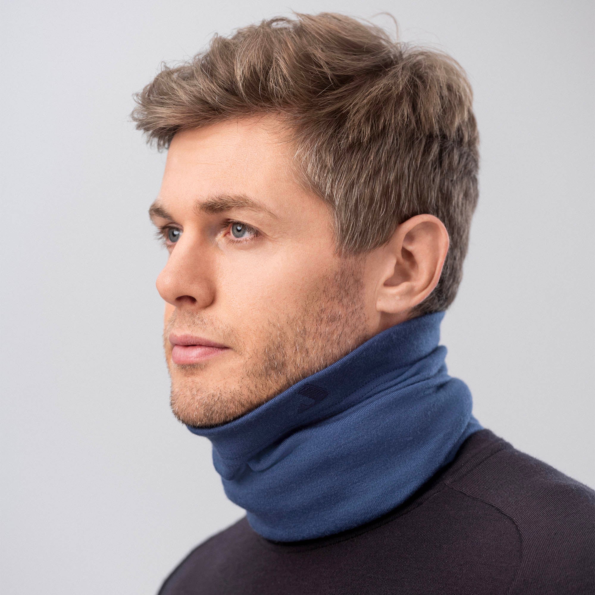 A man with short, light brown hair wears a black shirt and the Ensign Blue Neck Warmer Merino – AirWool 260, looking slightly left against a plain light gray background. This neck warmer provides warmth and breathability.
