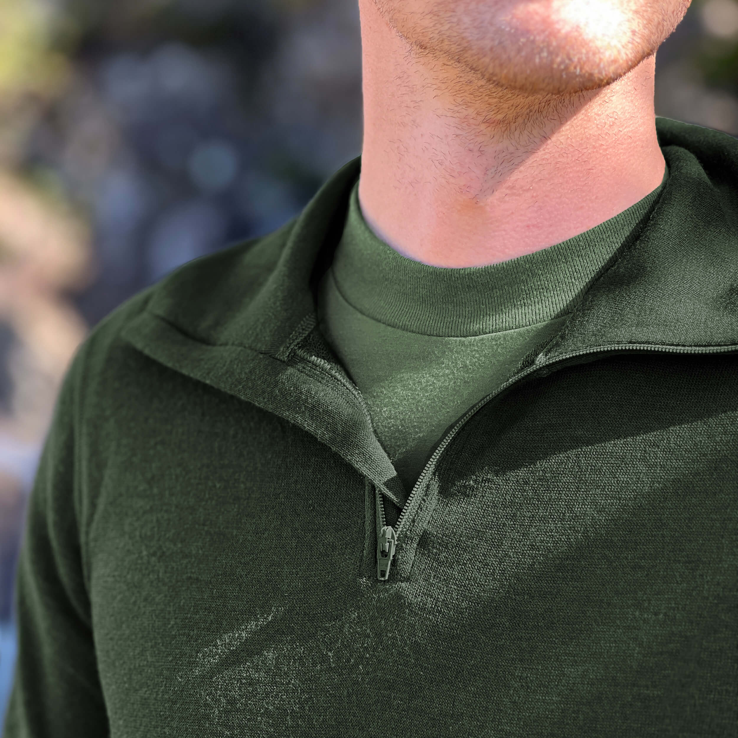 Close-up of a person wearing a Janus Norse Merino Polo in green, with a blurred natural background