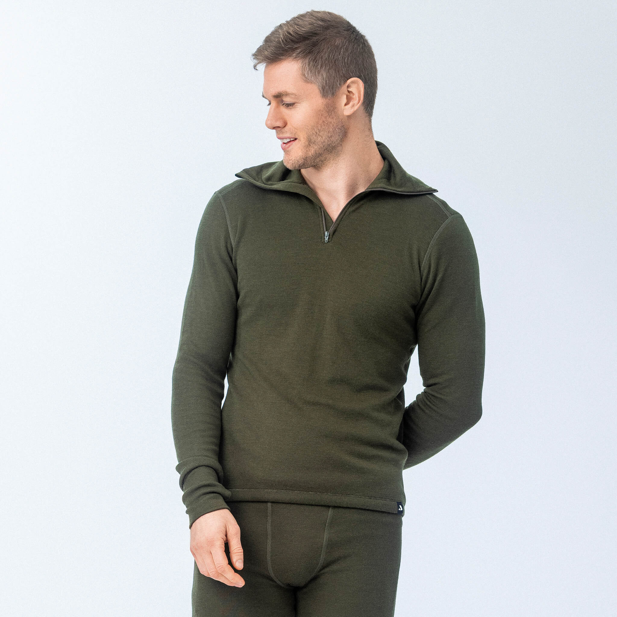 A man wears the Norse Merino 210 Unisex Polo with zip wool terry—an olive green, fitted, long-sleeve top and matching bottoms—standing against a plain backdrop. This versatile garment is perfect for outdoor adventures.