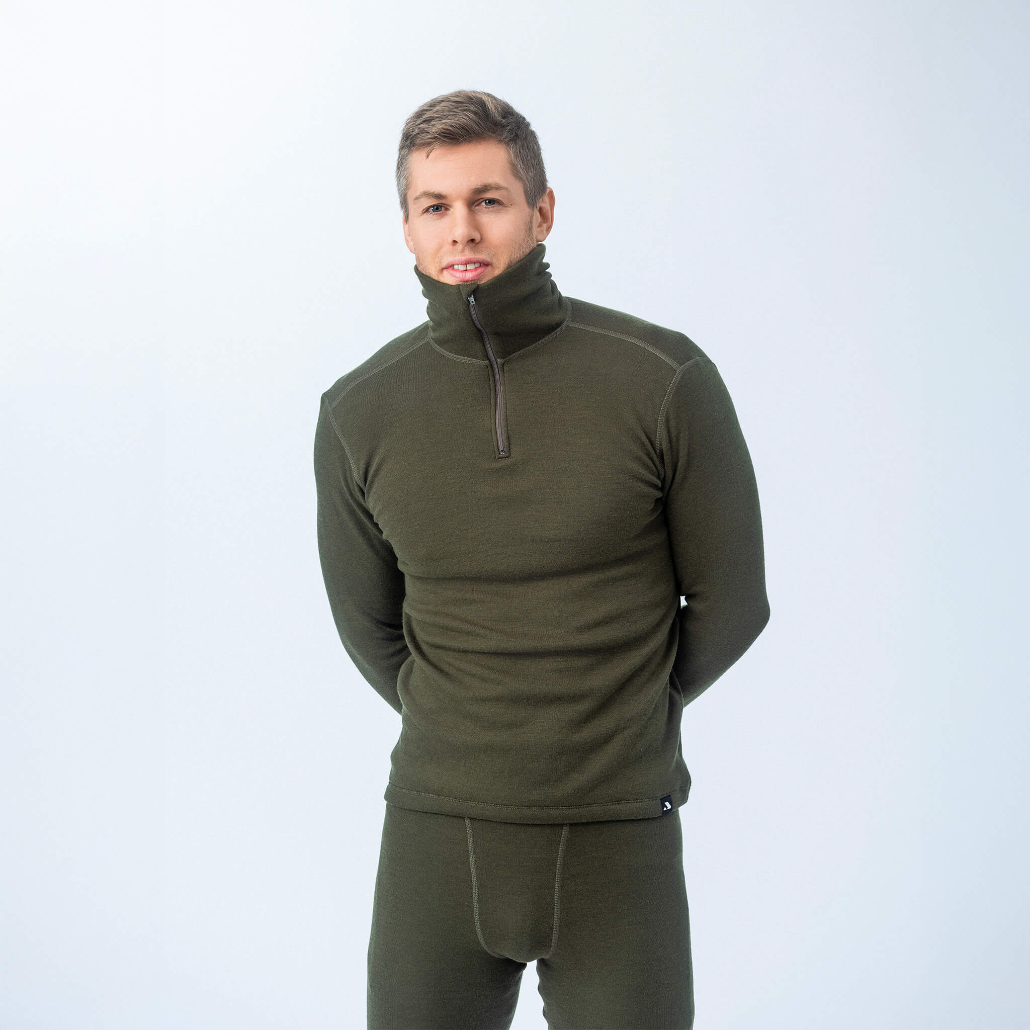A man stands against a plain light background wearing the Norse Merino 210 Unisex Polo with zip wool terry in green, featuring a high zippered collar—an ideal, versatile choice for outdoor activities.