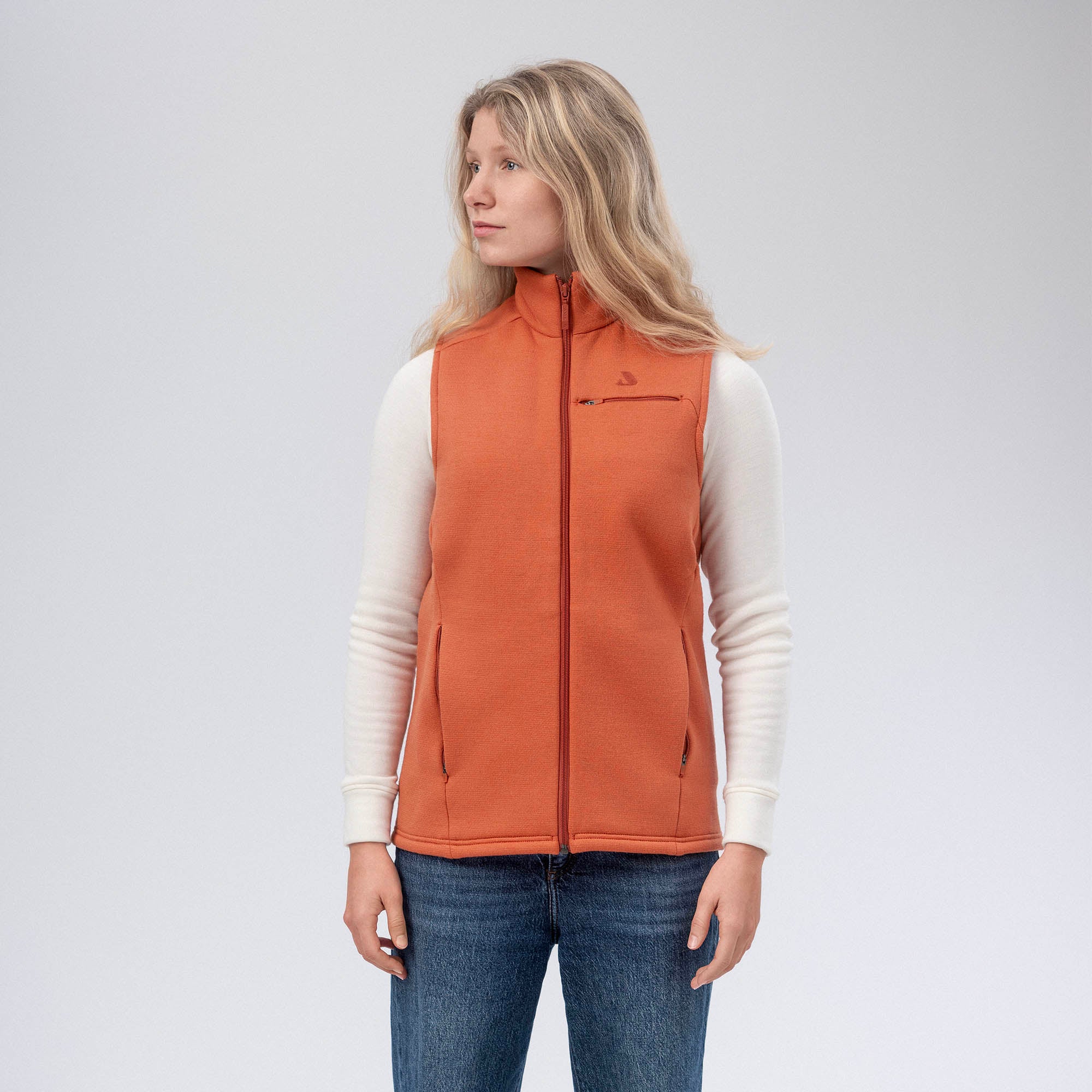 A woman with long blonde hair wears a white shirt, blue jeans, and the Vest Merino – AirWool 350 Women, known for warmth and breathability, standing against a plain light gray background and looking slightly to her left.