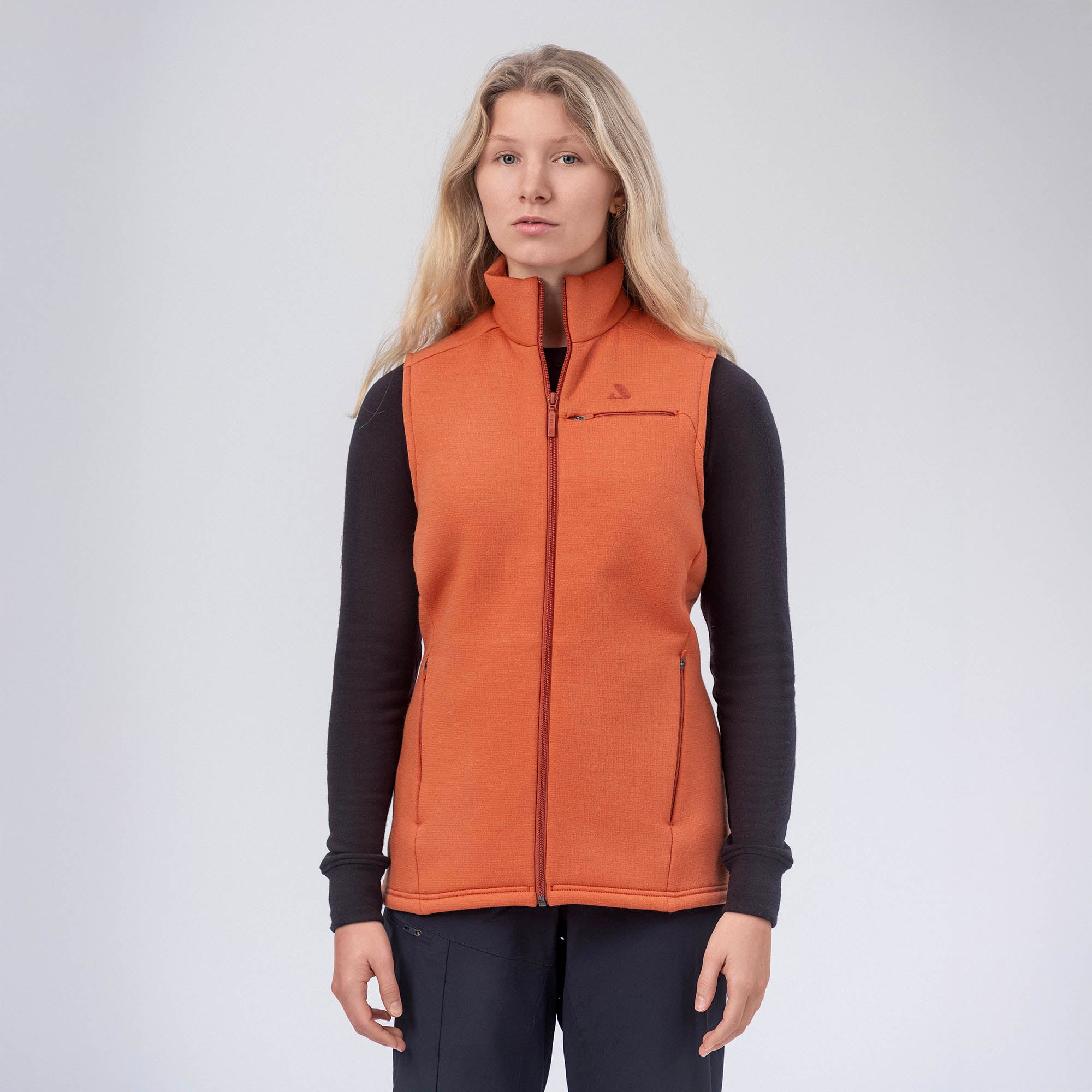 A woman with long blonde hair wears the Vest Merino – AirWool 350 Women in Mecca Orange over a black long-sleeve shirt and dark pants, standing against a plain light background, showcasing warmth and breathability.