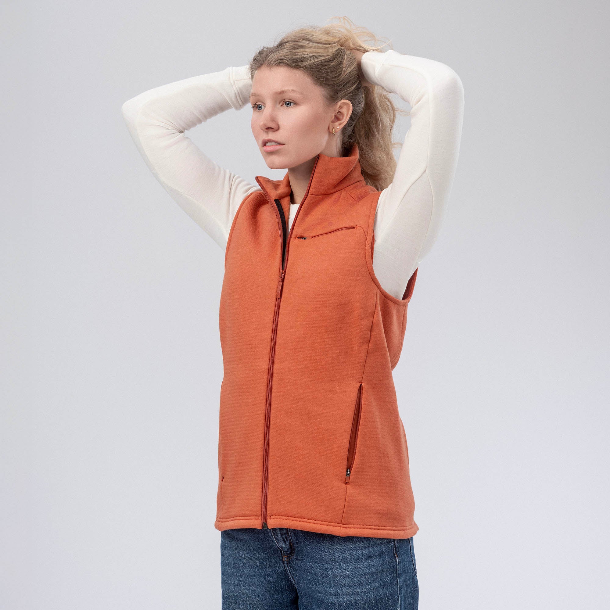 A woman in a white long-sleeve shirt, blue jeans, and the orange Vest Merino – AirWool 350 Women stands with hands behind her head against a plain background, highlighting the vest’s warmth and breathability.
