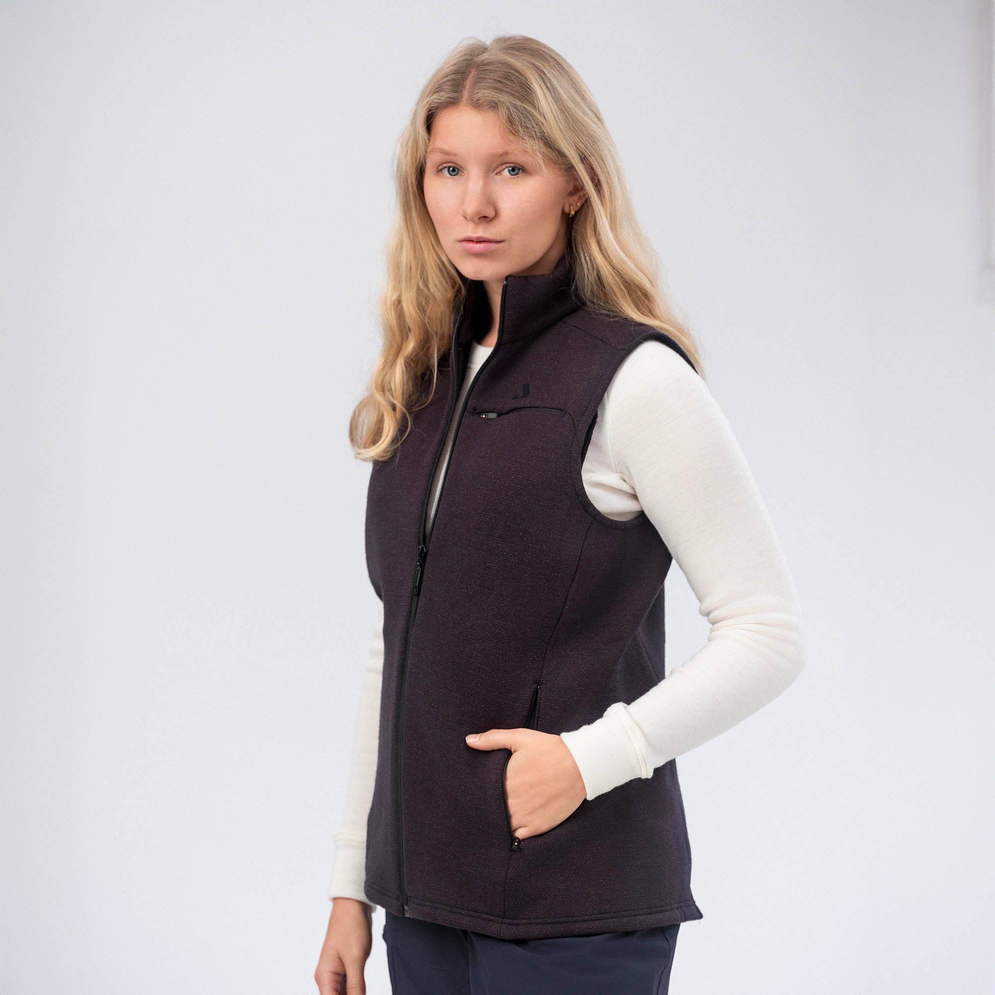 A woman with long blonde hair models the black Vest Merino – AirWool 350 Women over a white long-sleeve shirt, standing against a plain light background with one hand in her pocket, looking at the camera, highlighting warmth and breathability.