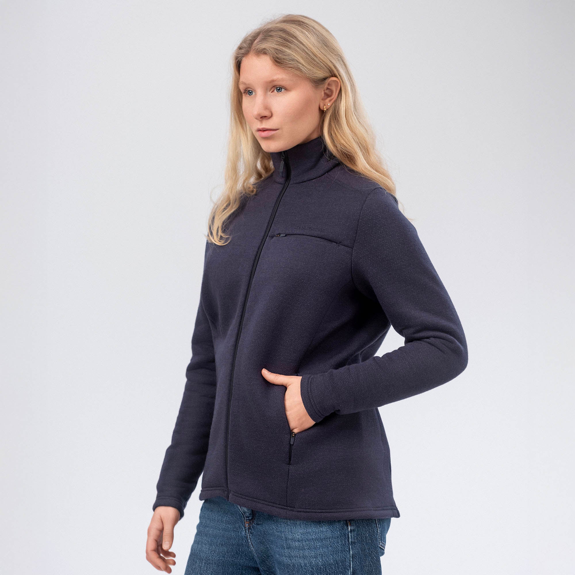 A woman with long blonde hair wears the Jacket Merino – AirWool 350 Women in navy and blue jeans, standing against a plain light background with one hand in her pocket, showcasing the jacket’s warmth and breathability.