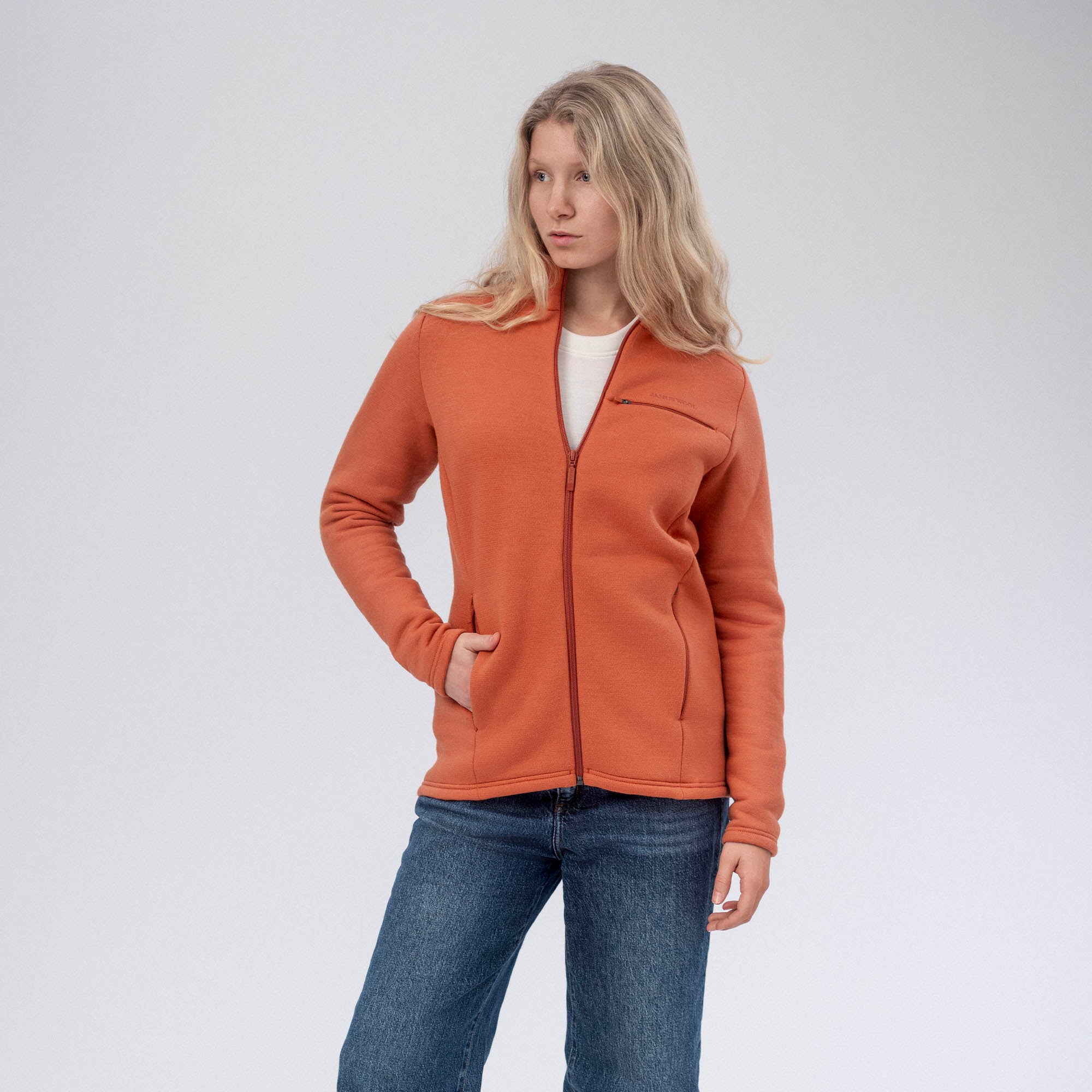 A woman with long blonde hair wears the Jacket Merino – AirWool 350 Women in Mecca Orange over a white shirt and blue jeans, standing against a plain light background with one hand in her pocket, highlighting the jacket’s breathability.