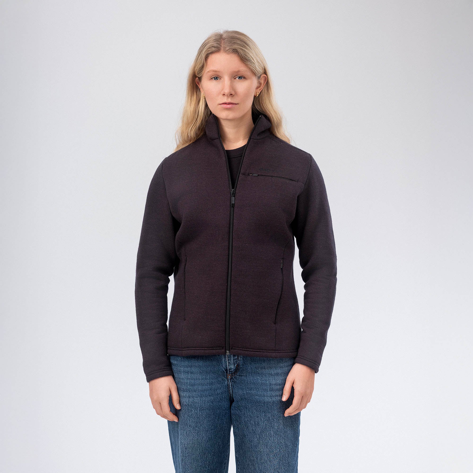 A woman with long blonde hair wears the black Jacket Merino – AirWool 350 Women, known for its breathability, and blue jeans as she stands facing forward against a plain white background.