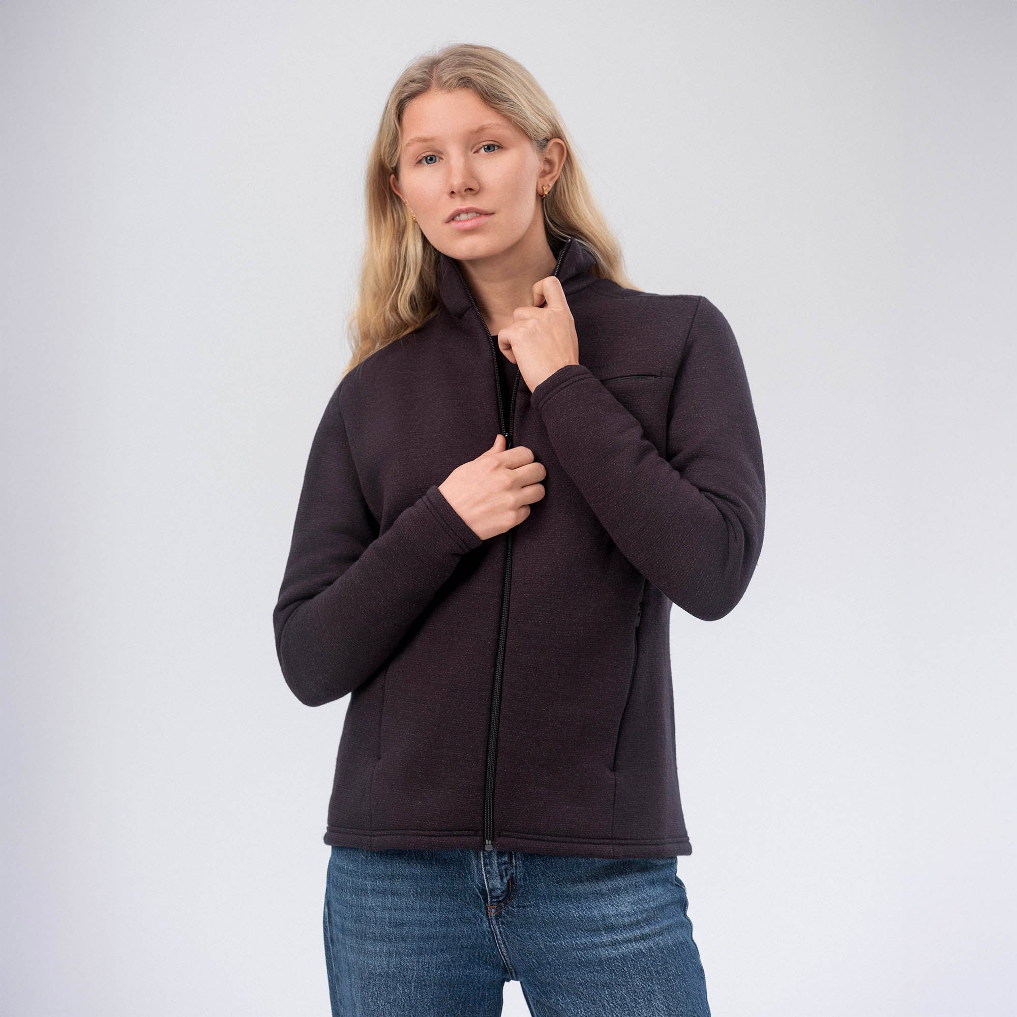 A woman with long blonde hair wears the Jacket Merino – AirWool 350 Women, known for breathability and warmth, over blue jeans. She stands against a plain light background, one hand pulling up the jacket zipper.
