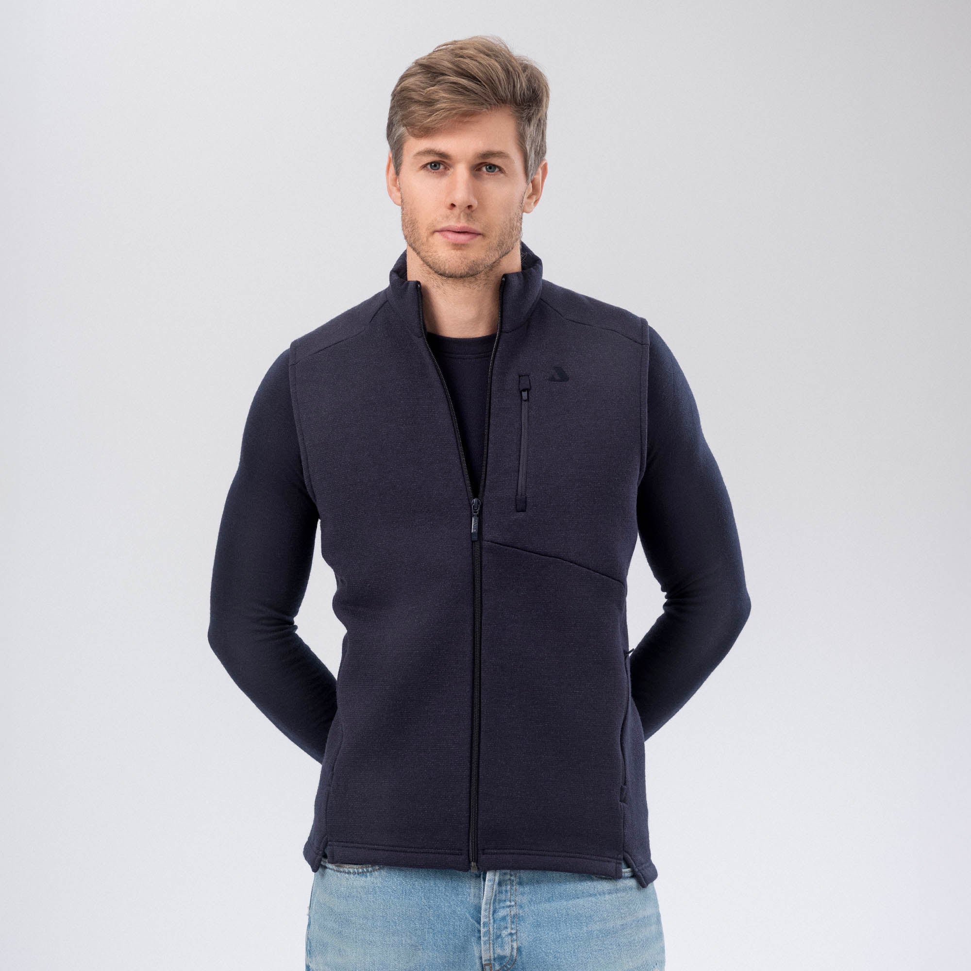 A man with light brown hair wears the Vest Merino – AirWool 350 Men in navy over a long-sleeve shirt and light blue jeans, standing against a plain light background, highlighting the vest’s warmth and breathability.