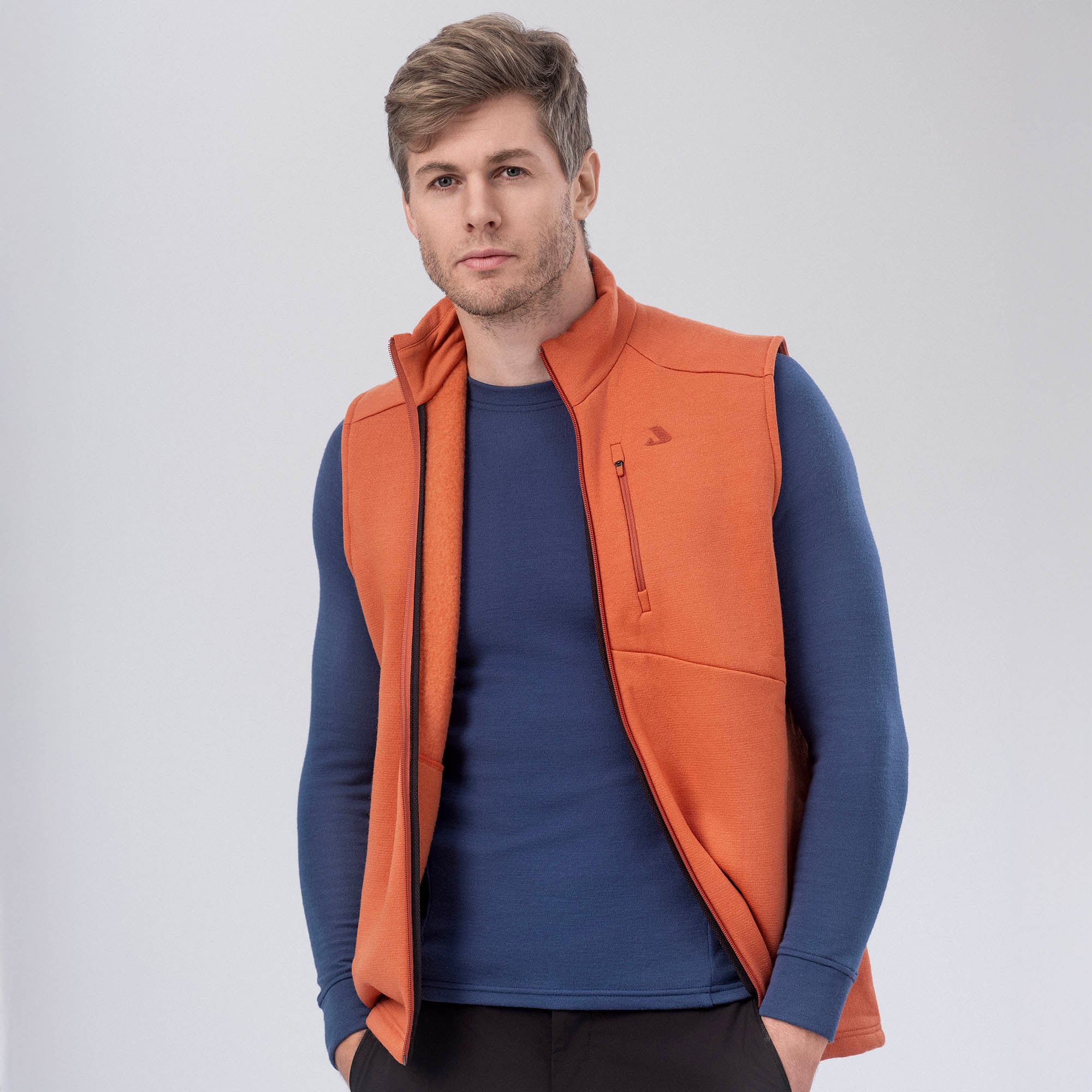 A man with light brown hair models the orange Vest Merino – AirWool 350 Men over a blue long-sleeve shirt, standing against a plain light gray background, highlighting the vest’s warmth and breathability.