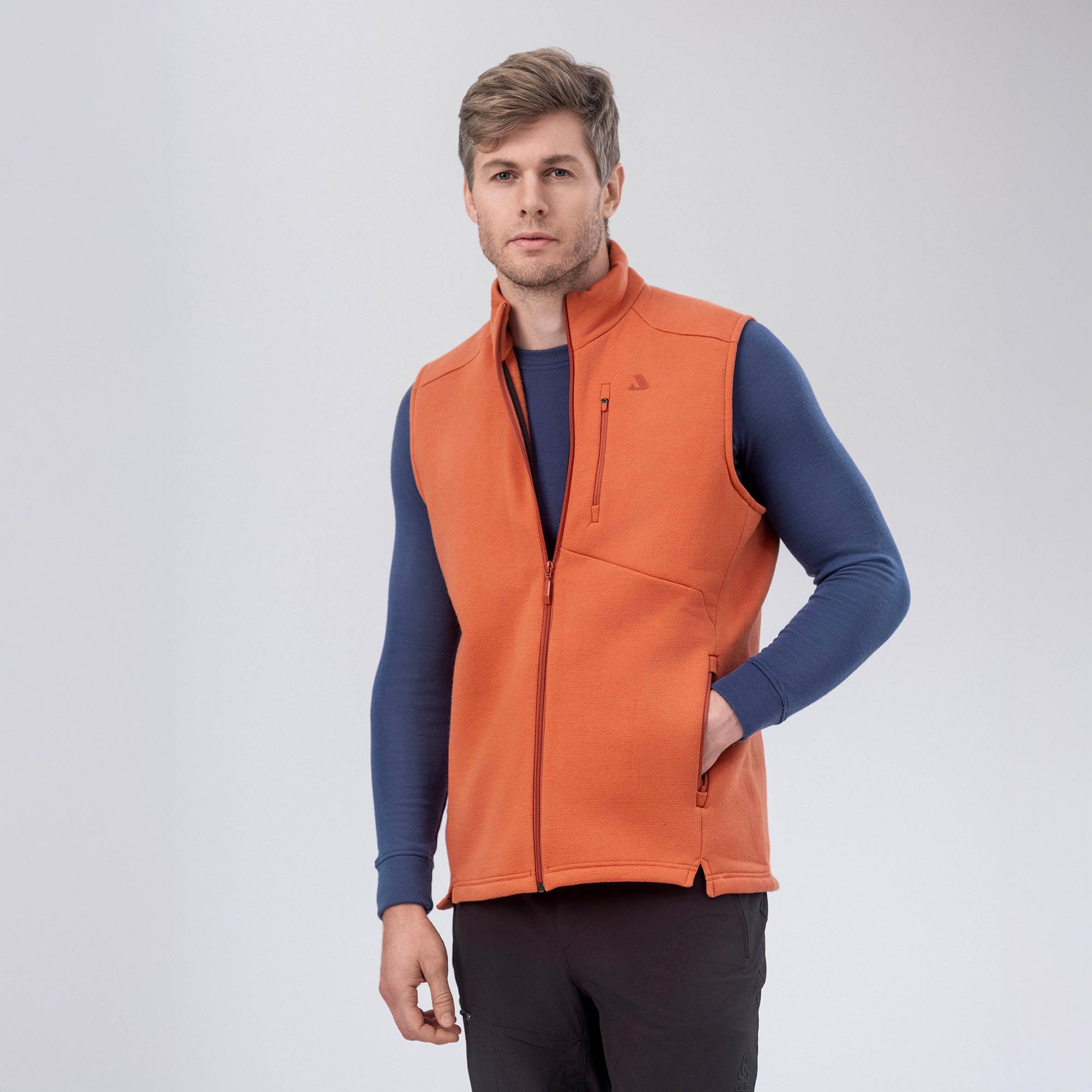 A man stands against a plain background wearing the orange Vest Merino – AirWool 350 Men over a long-sleeve blue shirt and black pants, one hand in his pocket, looking at the camera. The vest delivers warmth and breathability.