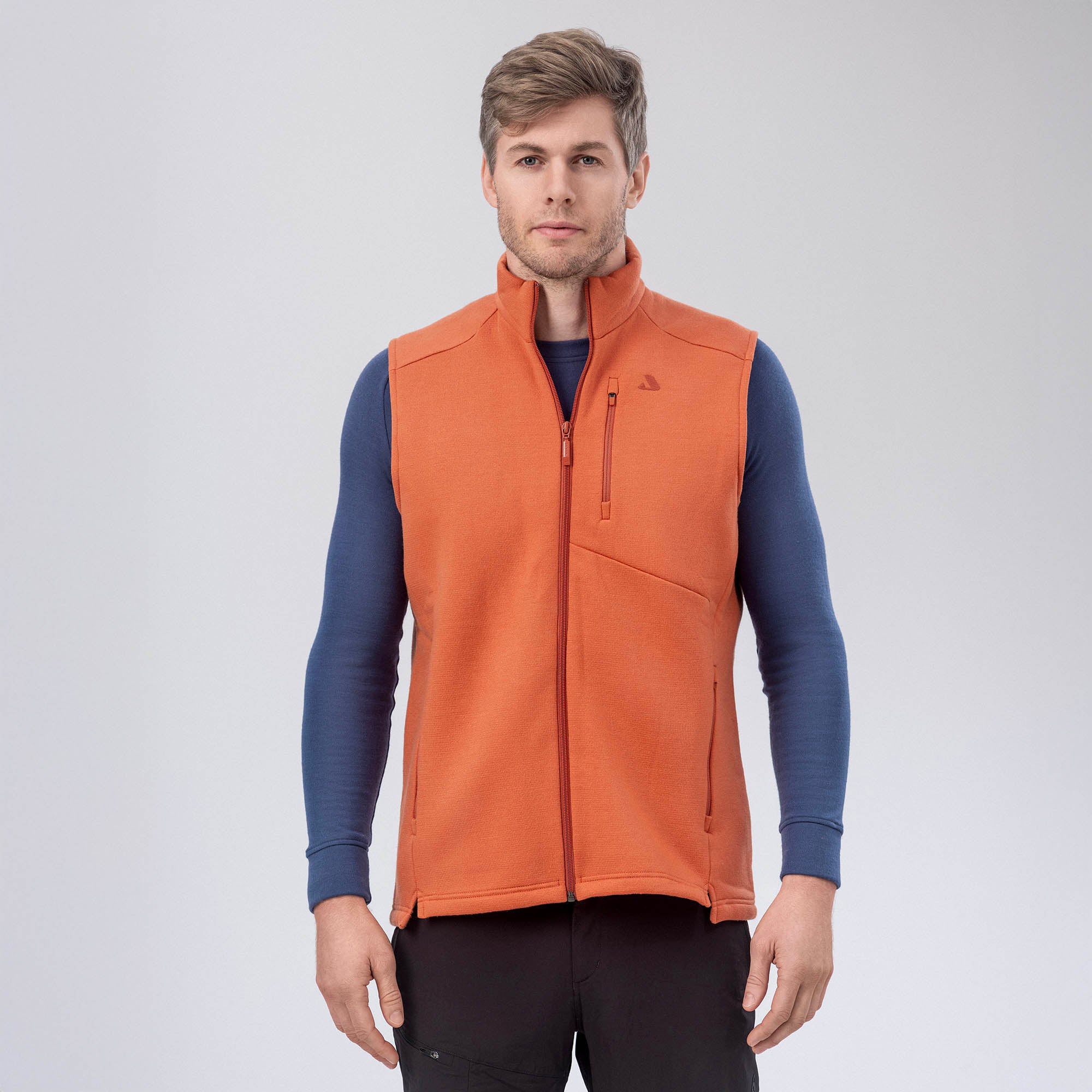 A man stands facing forward, wearing the Vest Merino – AirWool 350 Men in Mecca Orange over a blue long-sleeve shirt and black pants, set against a plain light gray background.