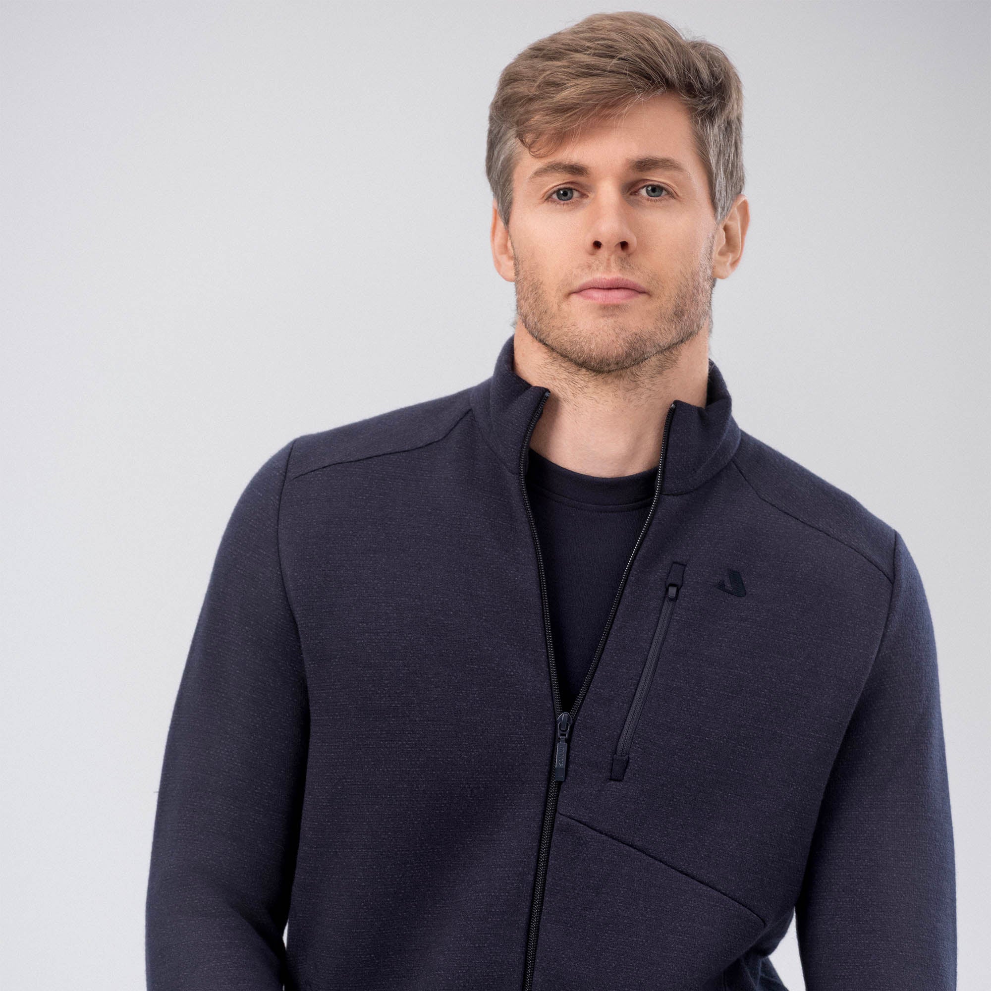 A man with light brown hair and a short beard models the Jacket Merino – AirWool 350 Men in navy, layered over a matching shirt, standing against a plain light gray background and gazing confidently at the camera.