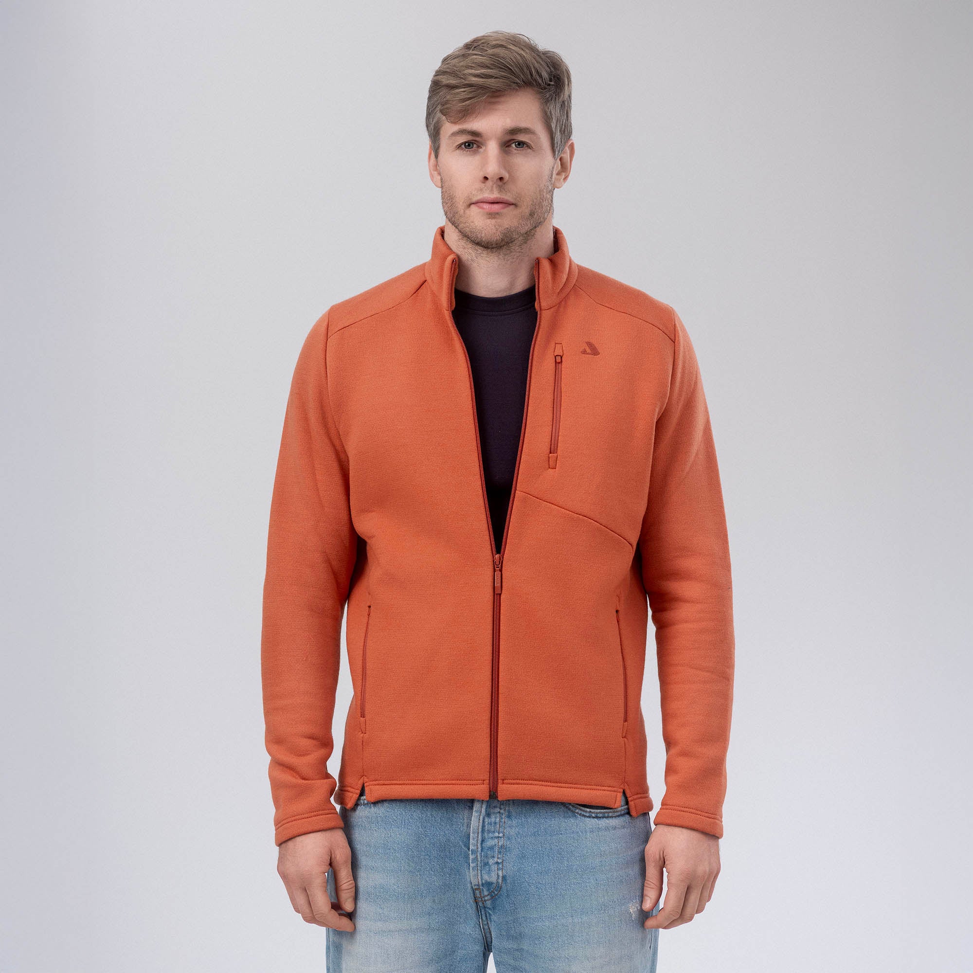 A man faces forward, wearing the burnt orange Jacket Merino – AirWool 350 Men over a dark shirt and light blue jeans, set against a plain light gray background.