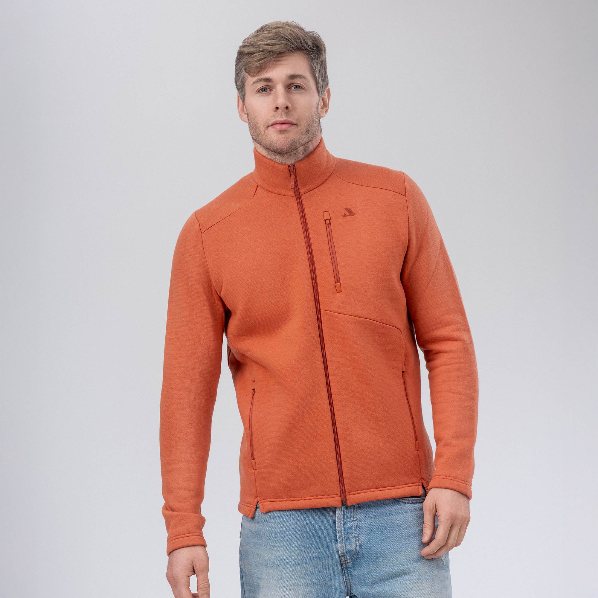 A man wearing the Jacket Merino – AirWool 350 Men in Mecca Orange with a high collar and light blue jeans stands against a plain light gray background.