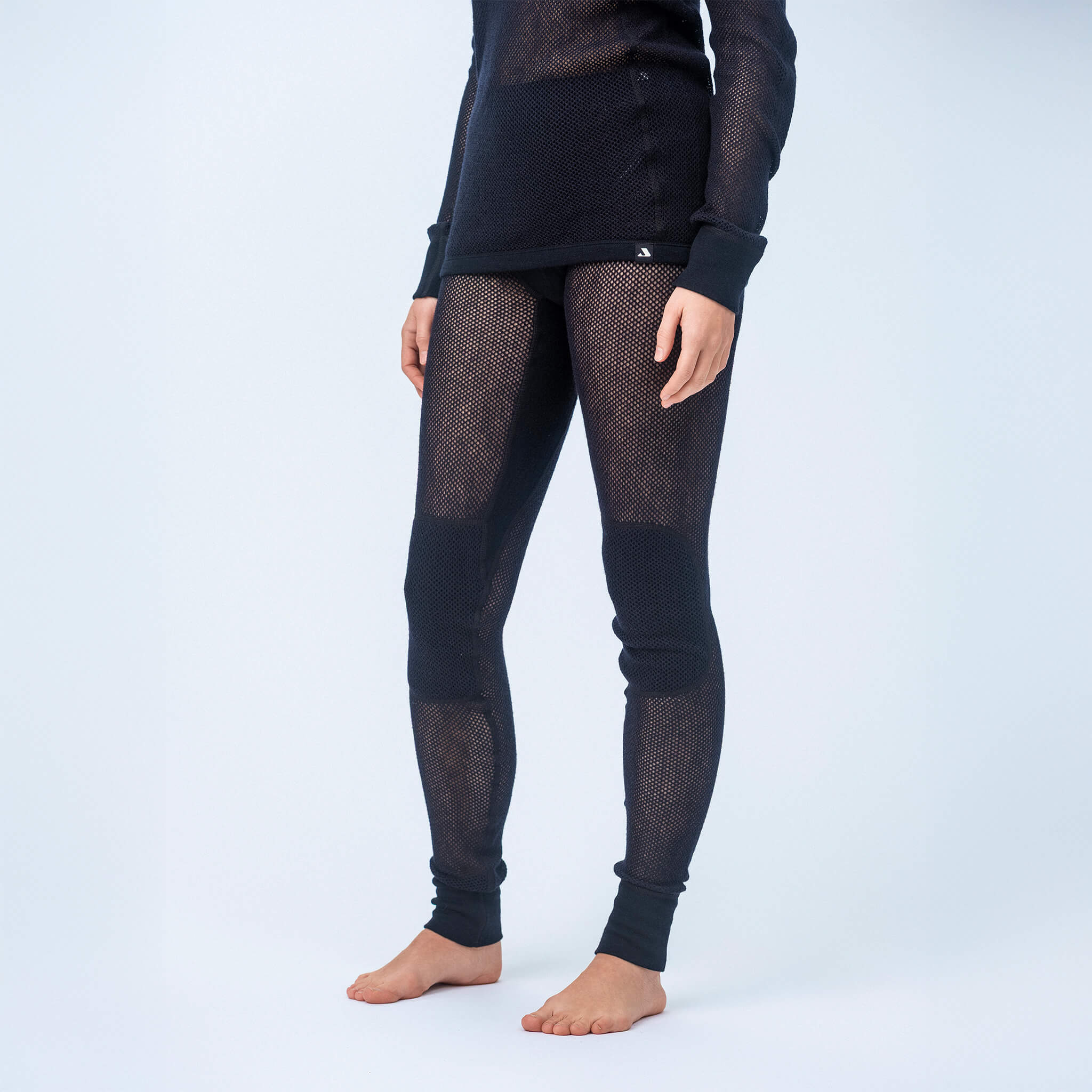 A person stands barefoot wearing black Norse Merino 165 Unisex mesh leggings and a matching wool mesh long-sleeve thermal top, set against a plain light background. The head and lower arms are out of frame.