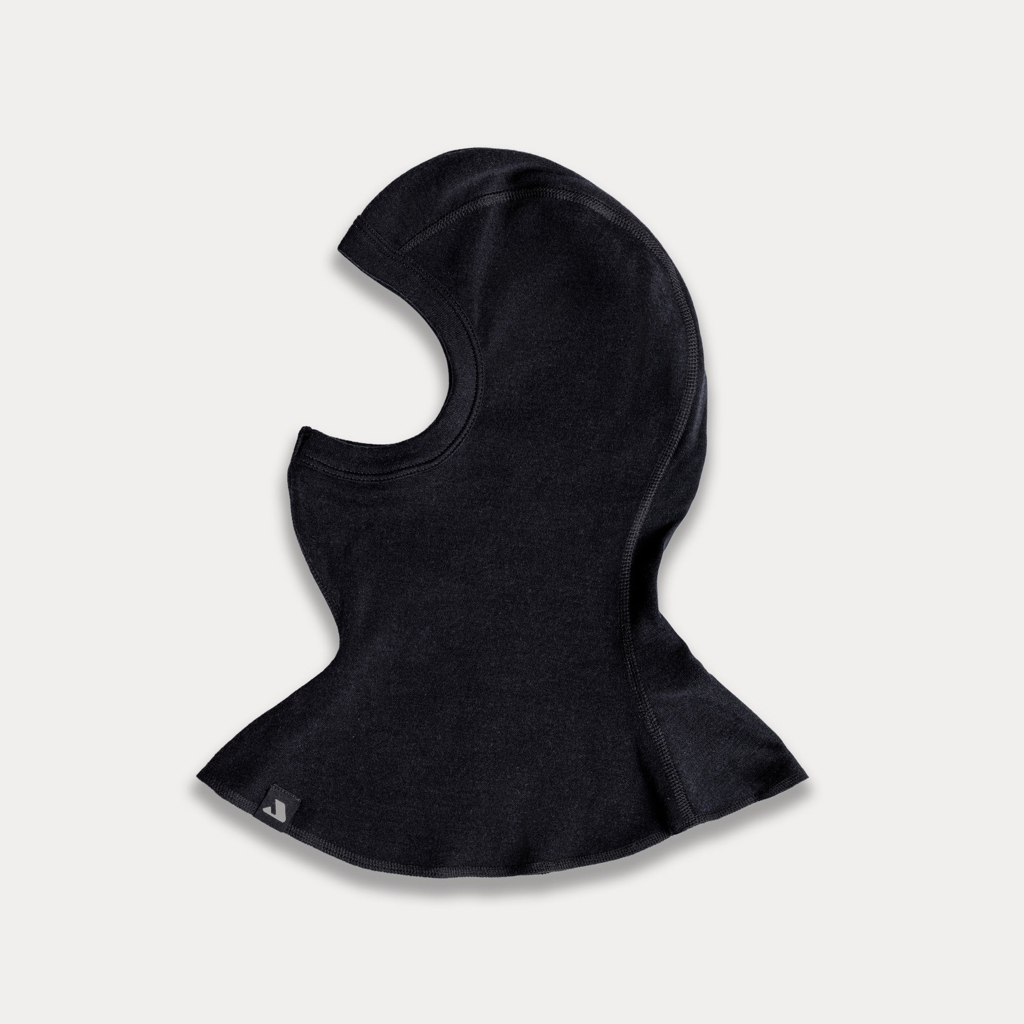 The Balaclava - Norse Merino 220 in black features an open face design and curved seams, made from fabric for warmth and cold protection. It flares at the bottom and is displayed flat on a light gray background.