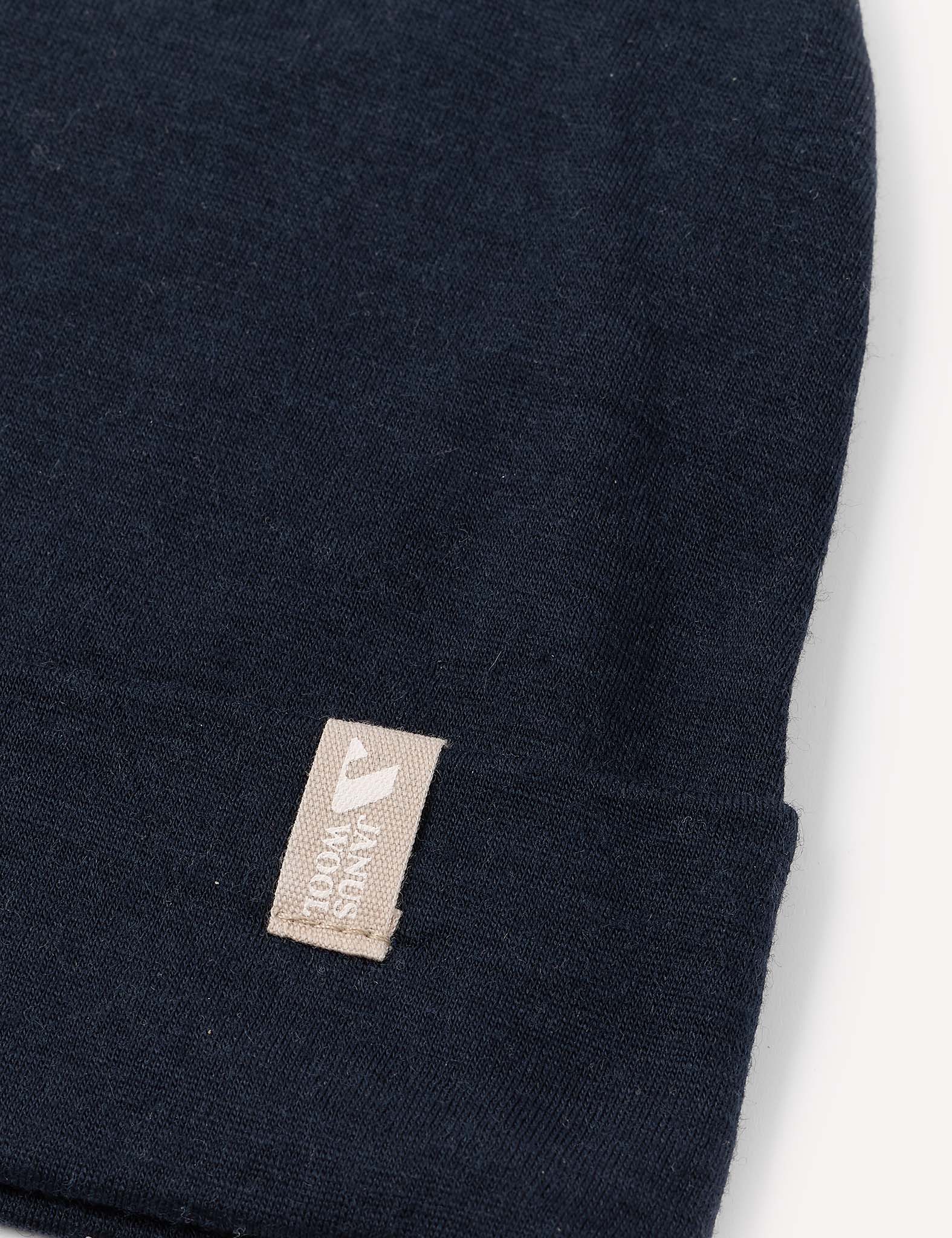 A close-up of the Beanie – Merino Kids Light in dark navy blue, featuring a small beige tag with partial white text near the folded edge. The background is plain white.