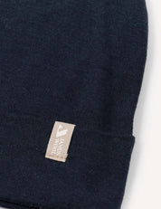 A close-up of the Beanie – Merino Kids Light in dark navy blue, featuring a small beige tag with partial white text near the folded edge. The background is plain white.
