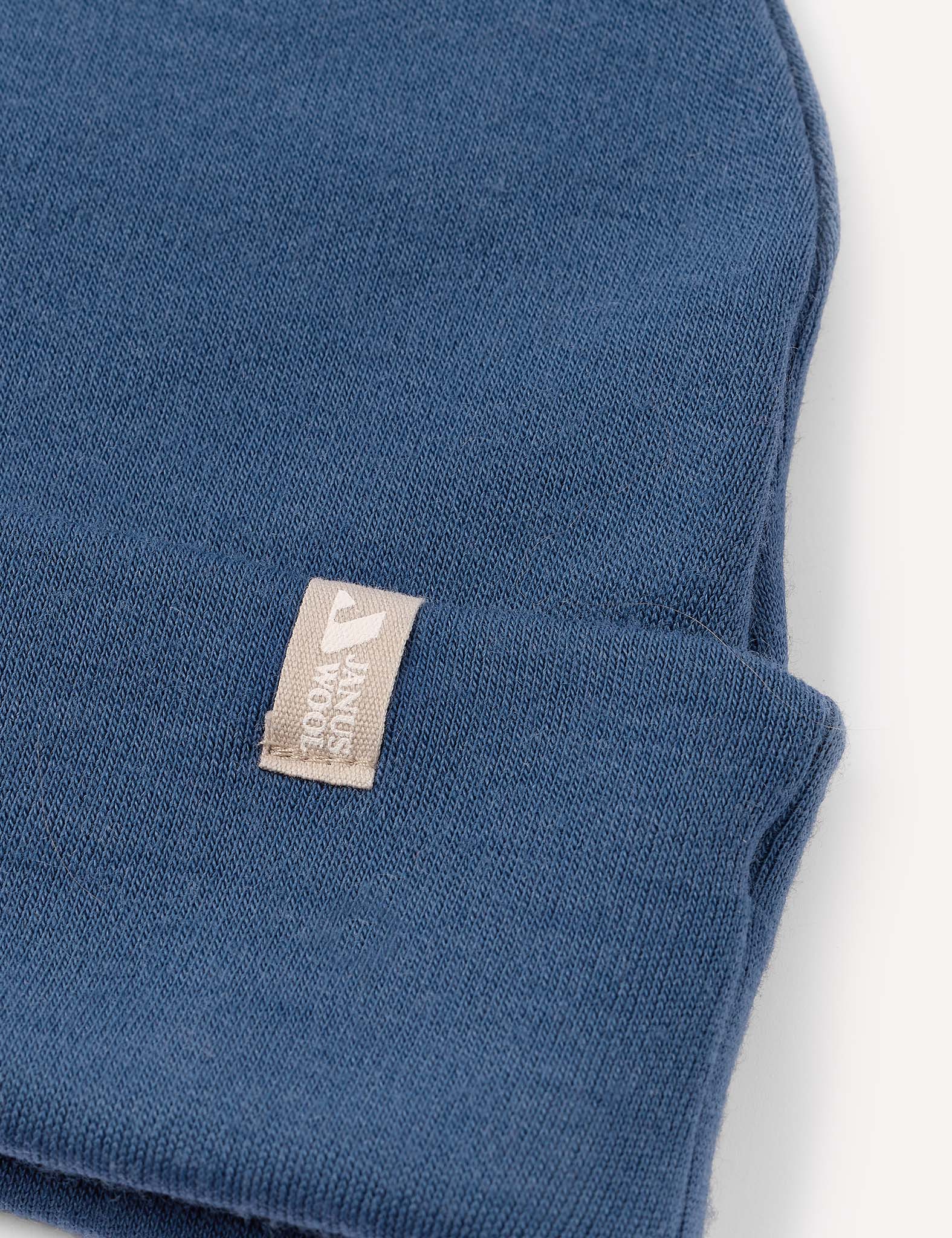 A close-up of the Beanie – Merino Kids AirWool in blue, featuring a beige tag with a white logo and text sewn onto the brim, set against a plain white background.