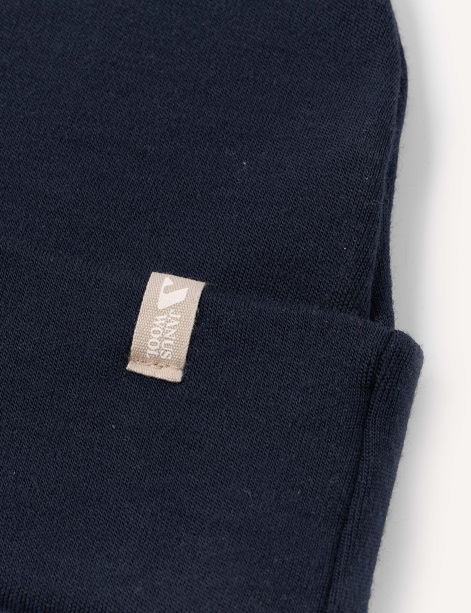 A close-up of the Beanie – Merino Kids AirWool in navy blue shows its soft, breathable merino wool fabric and a small beige tag with white text sewn onto the folded cuff.