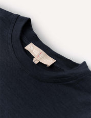 Close-up of the “Crewneck – Merino Kids Light,” a navy blue crew neck T-shirt made from soft merino wool jersey. A beige tag at the collar shows the brand details and care instructions. The shirt is laid flat on a light surface.