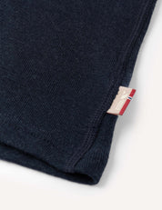 Close-up of the corner of the Crewneck – Merino Kids Classic Rib in dark blue ribbed wool jersey, featuring a small beige and red tag sewn into the seam, set against a plain off-white background.
